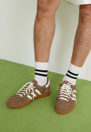 Man draagt bruine suède sneakers met witte strepen en witte sokken met twee zwarte strepen, staand op een groen tapijt.