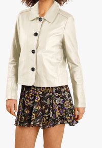 Ljust beige läderjacka med spetsig krage och svarta knappar, i kombination med en blomtryckt plisserad kjol i lila och gröna nyanser.