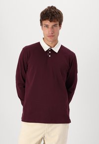 Polo shirt bordeaux a maniche lunghe con colletto bianco, tre bottoni e una texture del tessuto liscia. Vestibilità casual, design minimal.