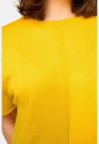 Haut jaune à manches courtes en tissu tricoté doux, avec un col rond et une couture visible au centre. Fini texturé et lisse.