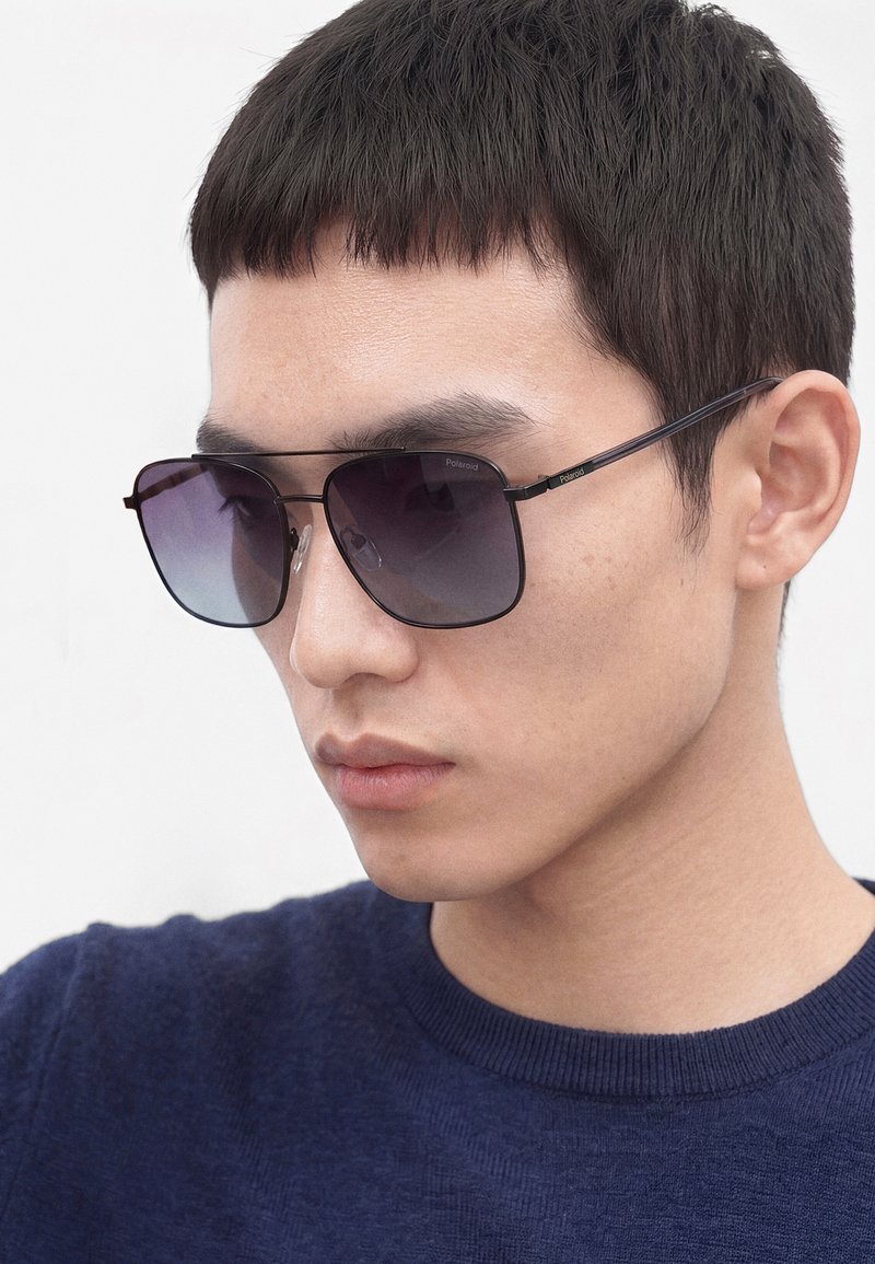 Jeune homme portant des lunettes de soleil carrées à dégradé foncé et un pull bleu marine, légèrement tourné vers la gauche sur un fond blanc.
