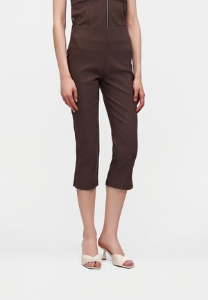 Modèle portant un pantalon court marron avec des détails de couture à l'avant, associé à des sandales blanches à talon ouvert, sur un fond uni.