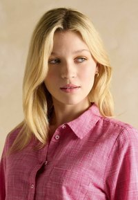 Svetloružová kockovaná košeľa s gombíkovým zapínaním, golierom a malou prednou vreckom. Tkanina sa zdá byť textúrovaná a priedušná.