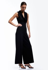 Schwarzer ärmelloser Jumpsuit mit drapiertem Ausschnitt und weitem Bein. Das Model trägt hochhackige Riemchensandalen. Glatte Stofftextur.