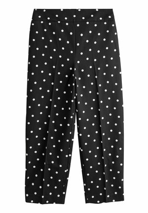 Close-up van zwarte broek en jas met witte stippen, hand die de zoom van de jas aanpast, draagt gouden armbanden om de pols.