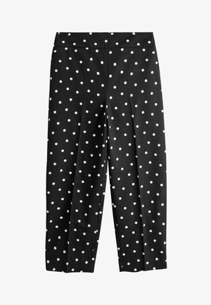 Close-up van zwarte broek en jas met witte stippen, hand die de zoom van de jas aanpast, draagt gouden armbanden om de pols.