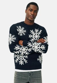 Maglione blu navy con motivi bianchi a fiocco di neve, texture lavorata a maglia, colletto rotondo e polsini a coste. Indossato sopra una camicia bianca.