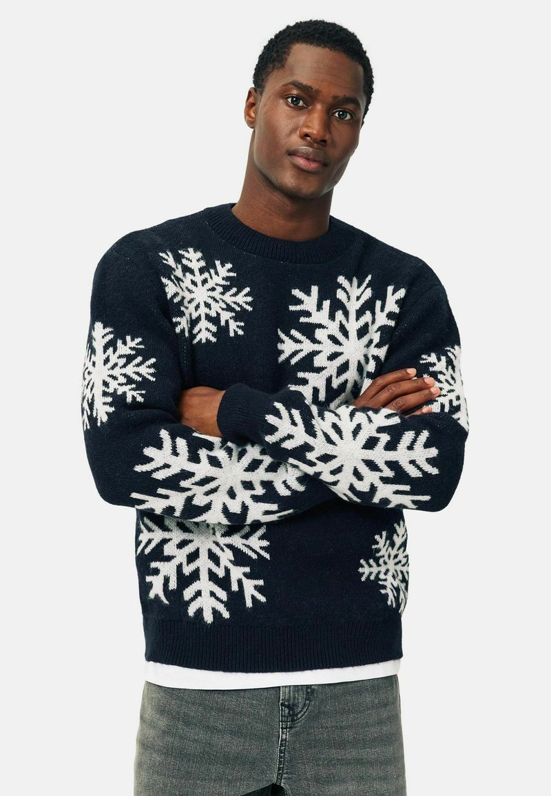 Maglione blu navy con motivi bianchi a fiocco di neve, texture lavorata a maglia, colletto rotondo e polsini a coste. Indossato sopra una camicia bianca.