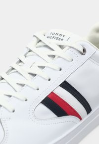 Baskets en cuir blanc avec des bandes en tissu noir et rouge, lacets blancs plats, et une étiquette logo sur la languette. Texture lisse, design minimaliste.