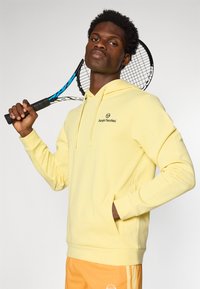 Gul hættetrøje med en frontlomme, snoretræk ved hætten og broderet logo. Modellen holder et tennisracket og viser dens sporty stil.