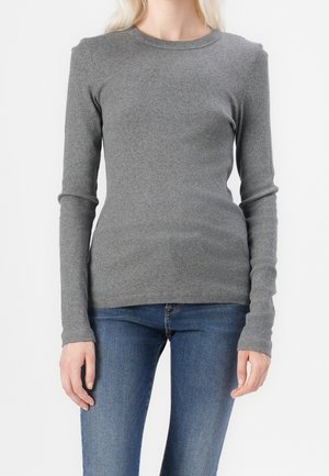 Woman wearing fitted long-sleeve gray sweater and blue jeans, standing against a plain white background.