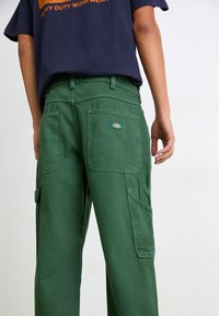 Green cargo pants made of heavy cotton, featuring multiple pockets and a textured finish. A logo patch is visible on the back pocket.