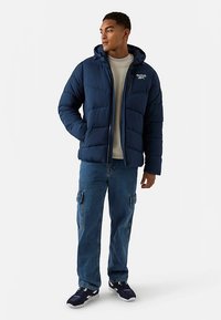 Chaqueta acolchada azul marino con capucha, cierre de cremallera y detalle de logotipo blanco, combinada con pantalones cargo azul claro y zapatillas oscuras.