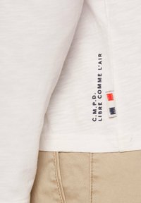 Hellbeiges Hemd aus einem strukturierten, weichen Stoff. Seitliches Etikett mit Text in Marineblau und Rot, der Marken- und Designdetails zeigt.