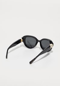 Ochelari de soare negri cu un cadru unghiular, lentile închise la culoare și accente aurii la balamale. Brațele sunt elegante și lucioase.