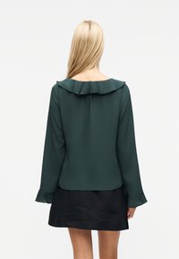 Ξανθιά γυναίκα με μακριά μαλλιά που φοράει μπλούζα μακρυμάνικη σκούρου πράσινου χρώματος με βολάν στον γιακά και στις μανσέτες και μαύρη φούστα, όρθια με την πλάτη προς τα εμπρός.