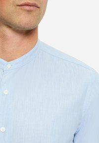 Himmelblaues Hemd mit Mandarin-Kragen und subtiler Textur, ausgestattet mit weißen Knöpfen und einem schlanken Schnitt, sichtbar am Halsausschnitt.