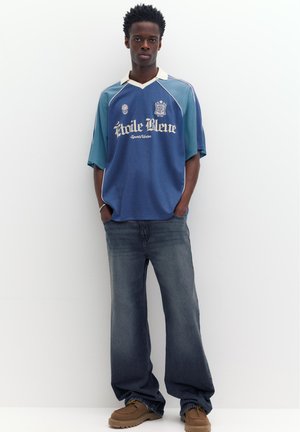 Ung mand står mod en hvid væg iført en oversized blå sportstrøje, løse mørke jeans og brune sko med hænderne i lommerne.