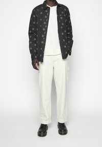 Zwart overhemd met patroon over een wit T-shirt, gecombineerd met crèmekleurige broek en zwarte schoenen. Het overhemd heeft een bloemenmotief in wit en gouden accenten.