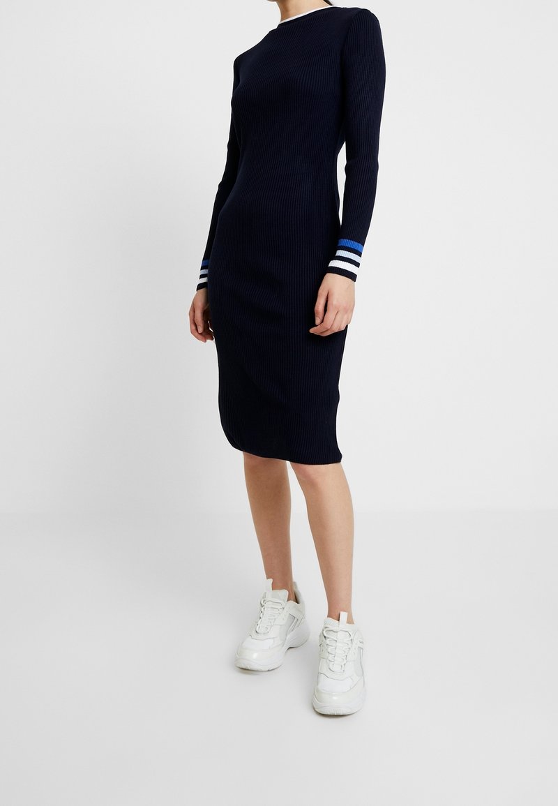Robe en maille côtelée navy à manches longues, comprenant des accents rayés bleus et blancs au niveau des poignets. Associée à des baskets de sport blanches.