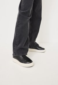 Črne usnjene superge z teksturirano površino in okroglo špicasto obliko, ki imajo ravne vezalke in belo gumijasto podplat, v kombinaciji s temno sivo jeans hlačami.