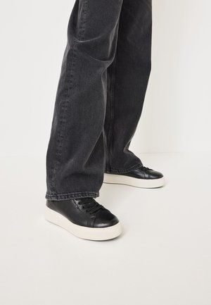 Black leather sneakers with a textured finish and round toe, featuring flat laces and a white rubber sole, paired with dark gray jeans.