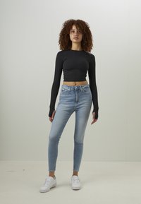 Μαύρο μακρυμάνικο crop top συνδυασμένο με ανοιχτό μπλε skinny jeans; Το μοντέλο φορά επίσης λευκά αθλητικά παπούτσια; Κομψός σχεδιασμός με εφαρμοστή σιλουέτα.