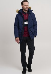 Mornarica izolirana parka s kapuco obloženo s krznom, ki ima dva sprednja žepa in zadrgo, oblečena čez maroon srajco z belimi črtami.