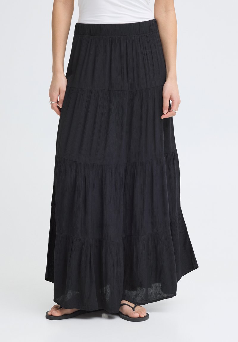 Femme portant une longue jupe noire à volants avec une ceinture élastique, associée à des tongs noires et un haut blanc partiellement hors cadre.