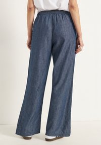 Denimbyxor med vida ben har ett elastiskt midjeband, blå färg och mjuk textur. Byxbenen smalnar av mot fållen, vilket ger en loose fit.