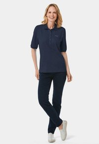 Navyblaues Poloshirt mit Knopfleiste, kurzen Ärmeln und Logodetails. Kombiniert mit dunklen Jeans und hellen Slippern.