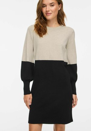 Femme portant une robe bicolore à manches longues avec le haut beige et le bas noir sur un fond blanc uni.