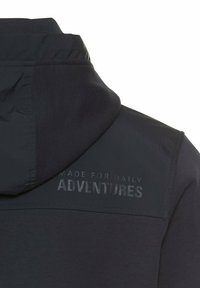 Mørk navy jakke med hætte, med en glat tekstur og en mat overflade. Tekst prægede på ryggen siger "LAVET TIL DAGLIGE EVENTYR."