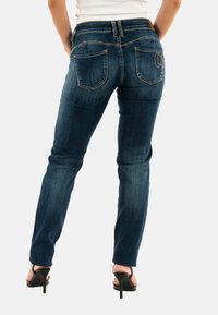 Jeans in denim blu con vestibilità slim, design a cinque tasche, dettagli cuciti e vita a media altezza. La vista posteriore mostra i dettagli distintivi sulle tasche.