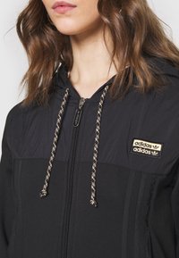 Giacca nera con zip e cappuccio, realizzata in tessuto testurizzato, con dettagli dorati e una patch con logo. I lacci di chiusura presentano un motivo intrecciato.