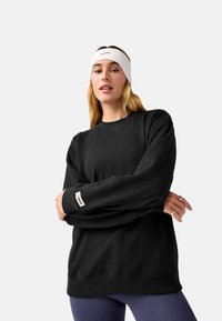 Felpa nera con collo a giro e polsini a coste, con etichetta del logo sulla manica. Il modello indossa una fascia bianca per capelli e leggings viola.