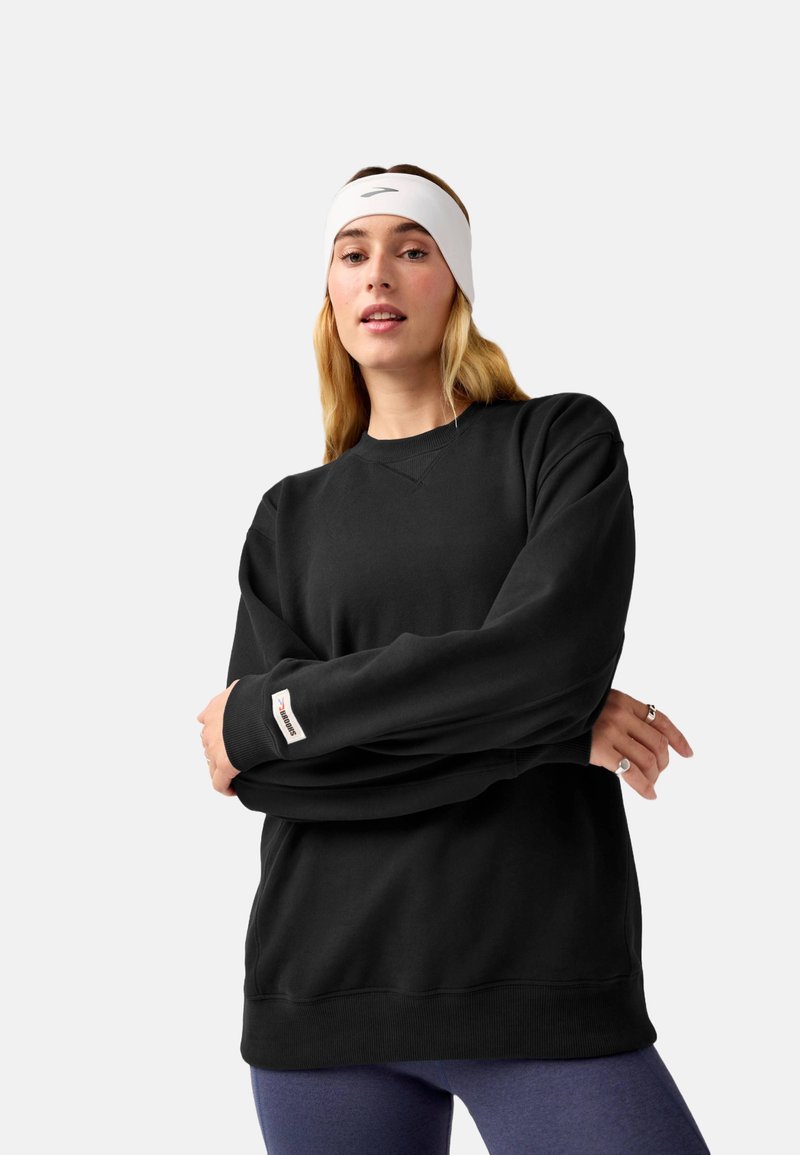 Felpa nera con collo a giro e polsini a coste, con etichetta del logo sulla manica. Il modello indossa una fascia bianca per capelli e leggings viola.