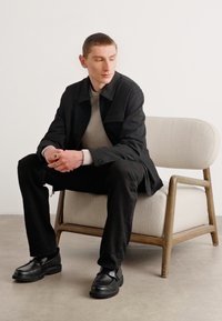 Black, textured jacket over a light grey top, paired with black pants and flat black shoes. Seated on a light, curved chair.