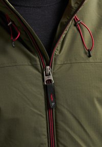 Grüne leichte Jacke mit Gittertextur, durchgehendem Reißverschluss mit roten Akzenten, verstellbaren Kordelzügen und schwarer Innenauskleidung.