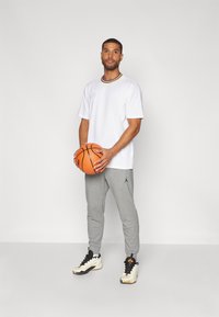 Hvit kortermet t-skjorte med lilla og gul krage, grå joggebukser, holder en oransje basketball. Svarte og hvite joggesko med detaljer.