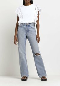 Vit bomullströja med volangärmar av öljett, tillsammans med ljusblå högmidjade utslitna raka jeans och svarta sandaler.