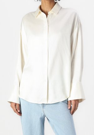Femme portant une chemise blanche ample à manches longues avec de larges poignets et un jean bleu clair sur un fond uni.