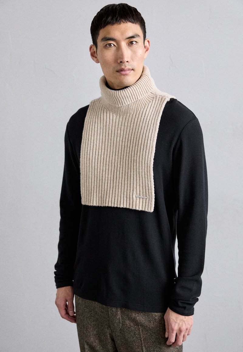 Μπεζ ριμπ τurtleneck κασκόλ πάνω από μαύρη μακρυμάνικη μπλούζα, με απαλή υφή και διακριτική λεπτομέρεια λογότυπου στην κάτω άκρη.