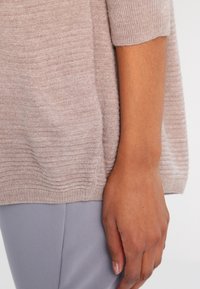 Pull tricoté beige clair à texture côtelée et manches courtes, associé à un pantalon gris doux. Mise au point sur les détails du tissu et de la couleur.