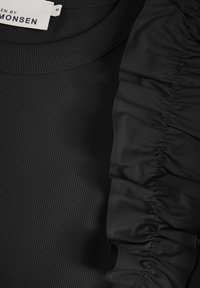 Haut noir à côtes avec des accents volantés sur une épaule, doté d'une encolure ronde et d'une texture douce. L'étiquette indique la marque et les informations de taille.