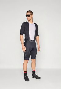 Vyriškas dviračių aprangos komplektas: trumpomis rankovėmis juoda marškinėliai ant baltos tinklelio pagrindo sluoksnio, derinami su prigludusiais tamsiais šortais. Juodi dviratininko batai ir kojinės.