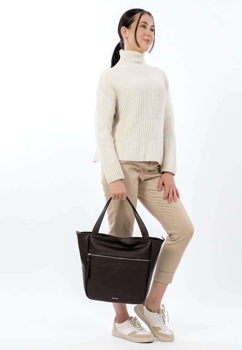 Bruine leren shopper met ritssluiting, dubbele stevige handvatten en een gestructureerd oppervlak. Draagt beige broek en casual sneakers.