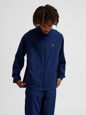 Mand iført en navyblå jakke med lynlås og matchende bukser, kigger ned med hænderne nær jakkelommerne foran en ensfarvet baggrund.