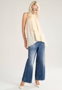 Top senza maniche color crema con balze sovrapposte, abbinato a jeans a gamba larga blu sbiaditi e décolleté bianchi a punta.