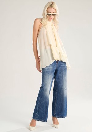 Top senza maniche color crema con balze sovrapposte, abbinato a jeans a gamba larga blu sbiaditi e décolleté bianchi a punta.
